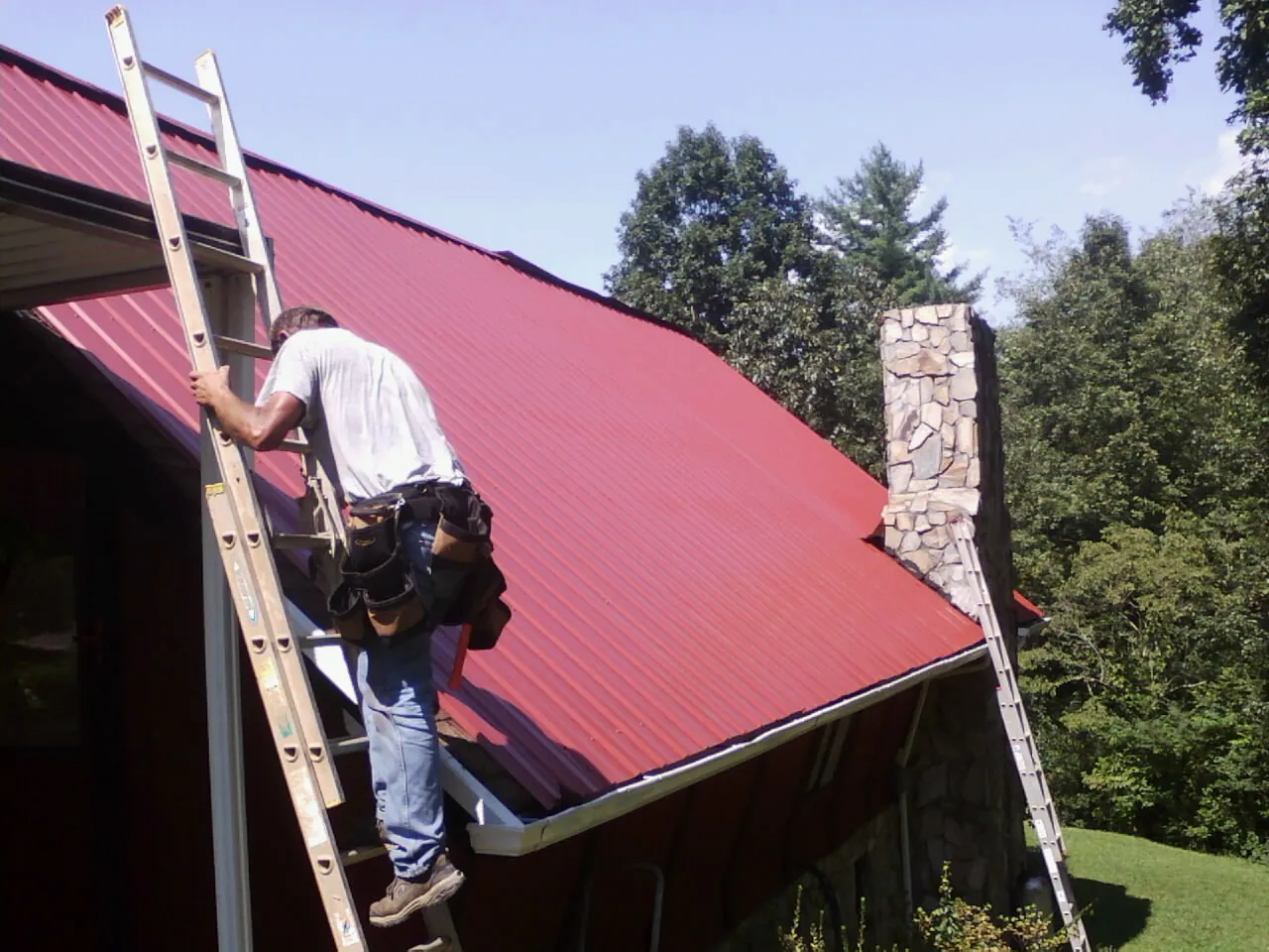Roofer on a ladder beside a red metal roof and stone chimney — Soffit & Fascia Repair by Roofing Brothers in Galesville